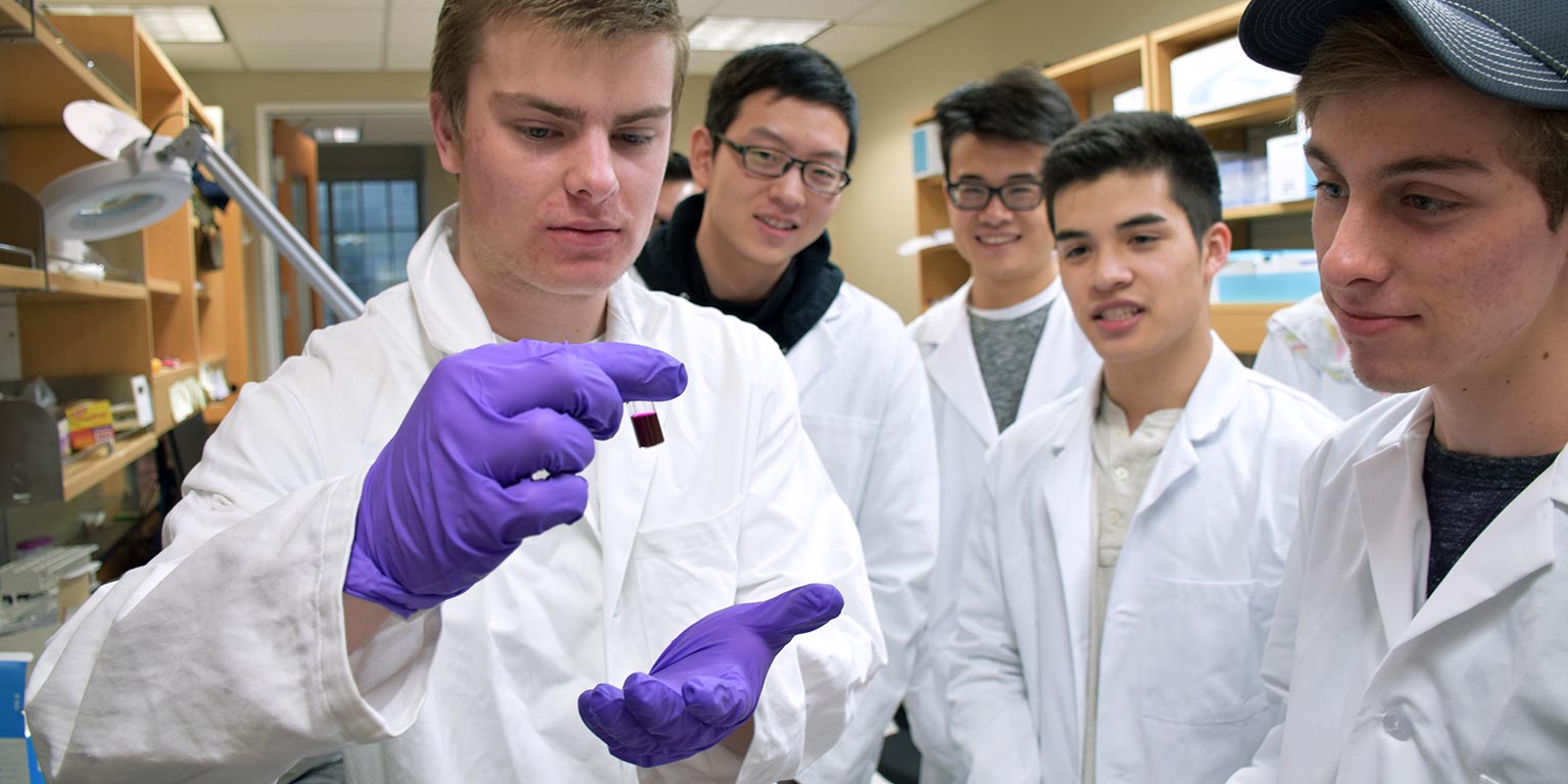 Students in white lab coats examine a sample in a test tube.