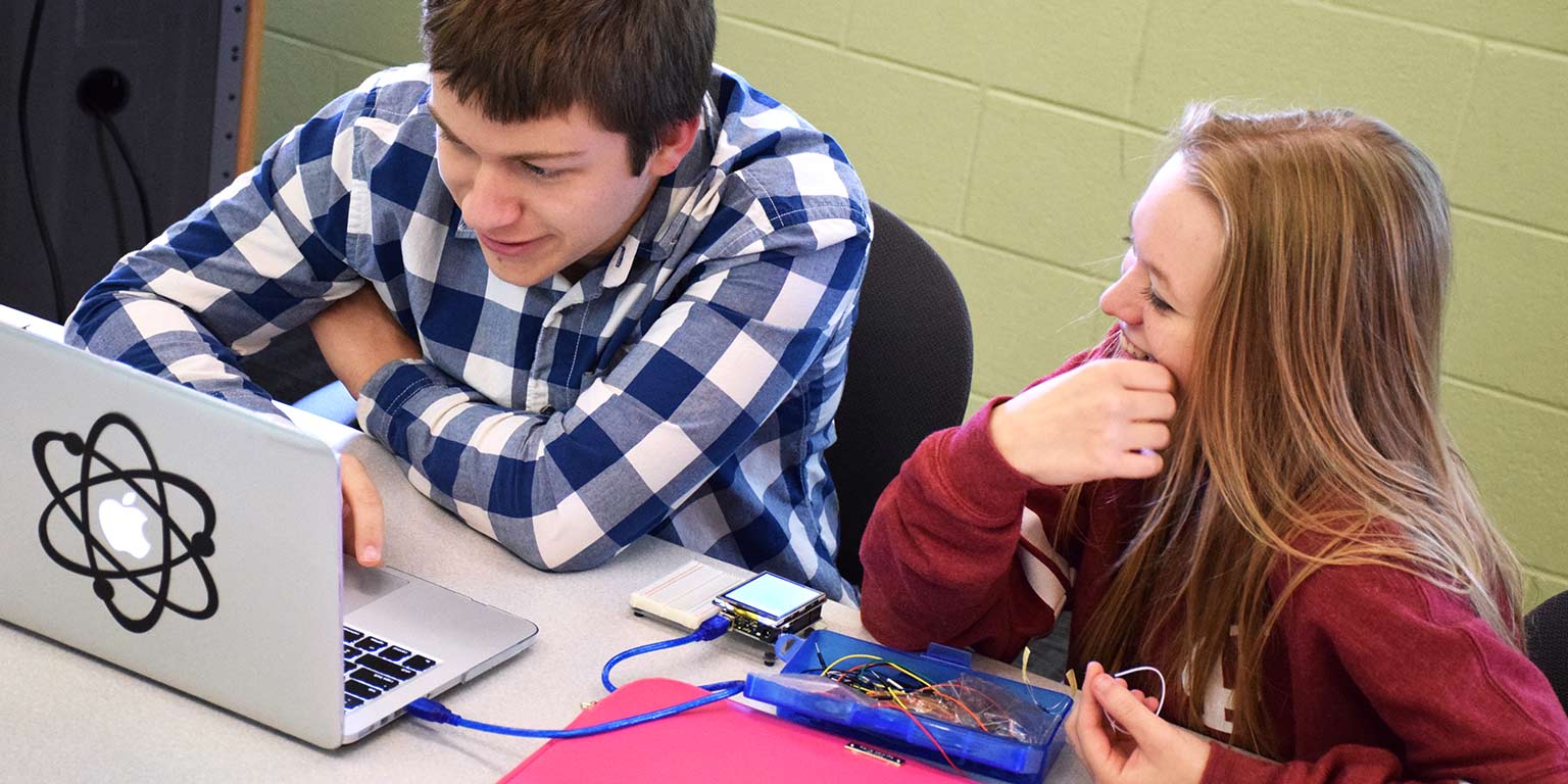 Two students look at a laptop that's hooked up to a project.